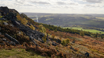 Fern covered hillside This landscape photograph captures a fern covered hillside along Curbar Edge in Derbyshire, United Kingdom, within the Peak District National Park. Taken in the early afternoon during autumn, the image showcases a rugged scene of nature with rocky outcrops and vibrant autumnal vegetation. The rural countryside stretches out into the distance, revealing rolling farmland and wooded areas typical of the Peak District. In the background, the elevated vantage point provides a wide view of the valley below, with fields patchworked by hedgerows and scattered trees beneath a partly cloudy sky. The image highlights the natural beauty and distinctive geological features of Curbar Edge, illustrating the landscape typical of rural Derbyshire in the autumn season.